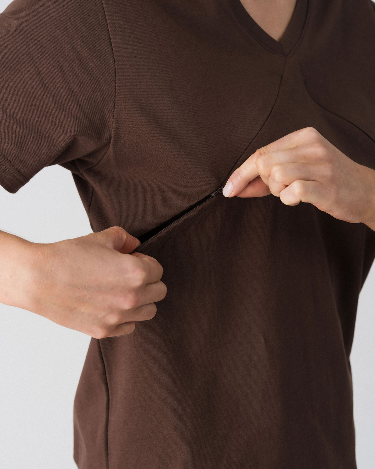 Model opening the hidden diagonal zip below the V-neckline of the Lenelly Avery tee in brown for discreet breastfeeding access