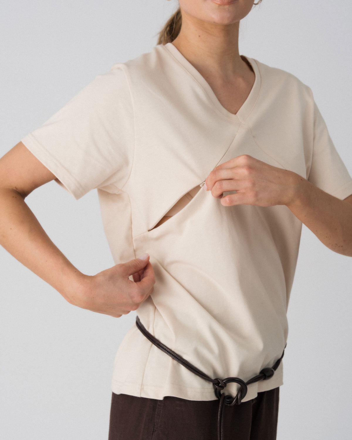 Model opening the hidden diagonal zip below the V-neckline of the Lenelly Avery tee in sand for discreet breastfeeding access