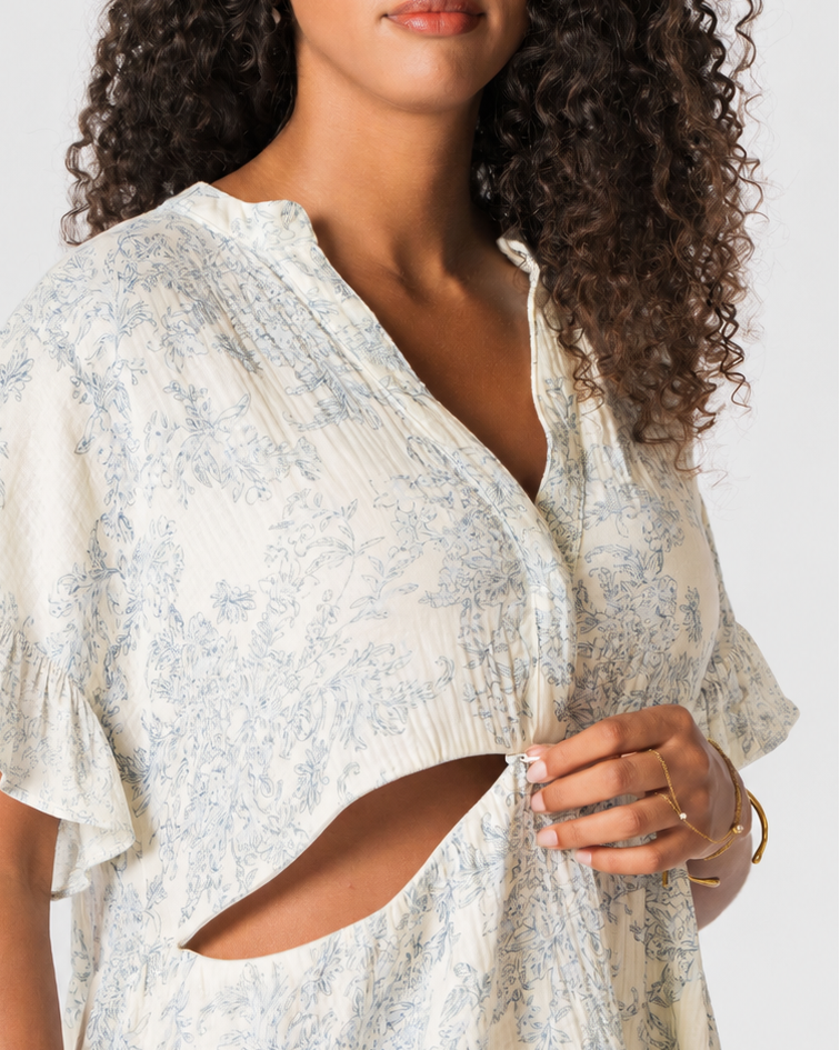 A woman wearing a light cream-colored short-sleeve tunic dress with a blue floral toile pattern, featuring a V-neckline and a distinctive side cutout, photographed against a neutral light gray background.