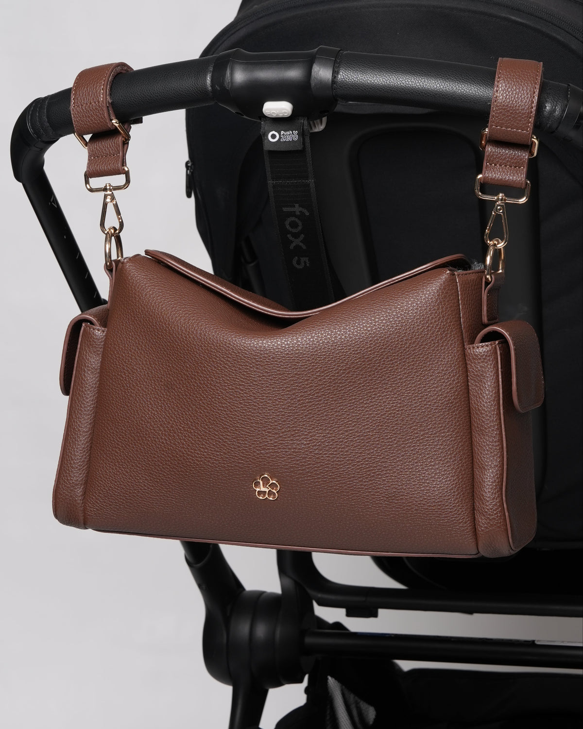 A brown pebbled leather shoulder bag with gold hardware and a small gold logo, hanging from a black stroller handlebar against a light gray background.