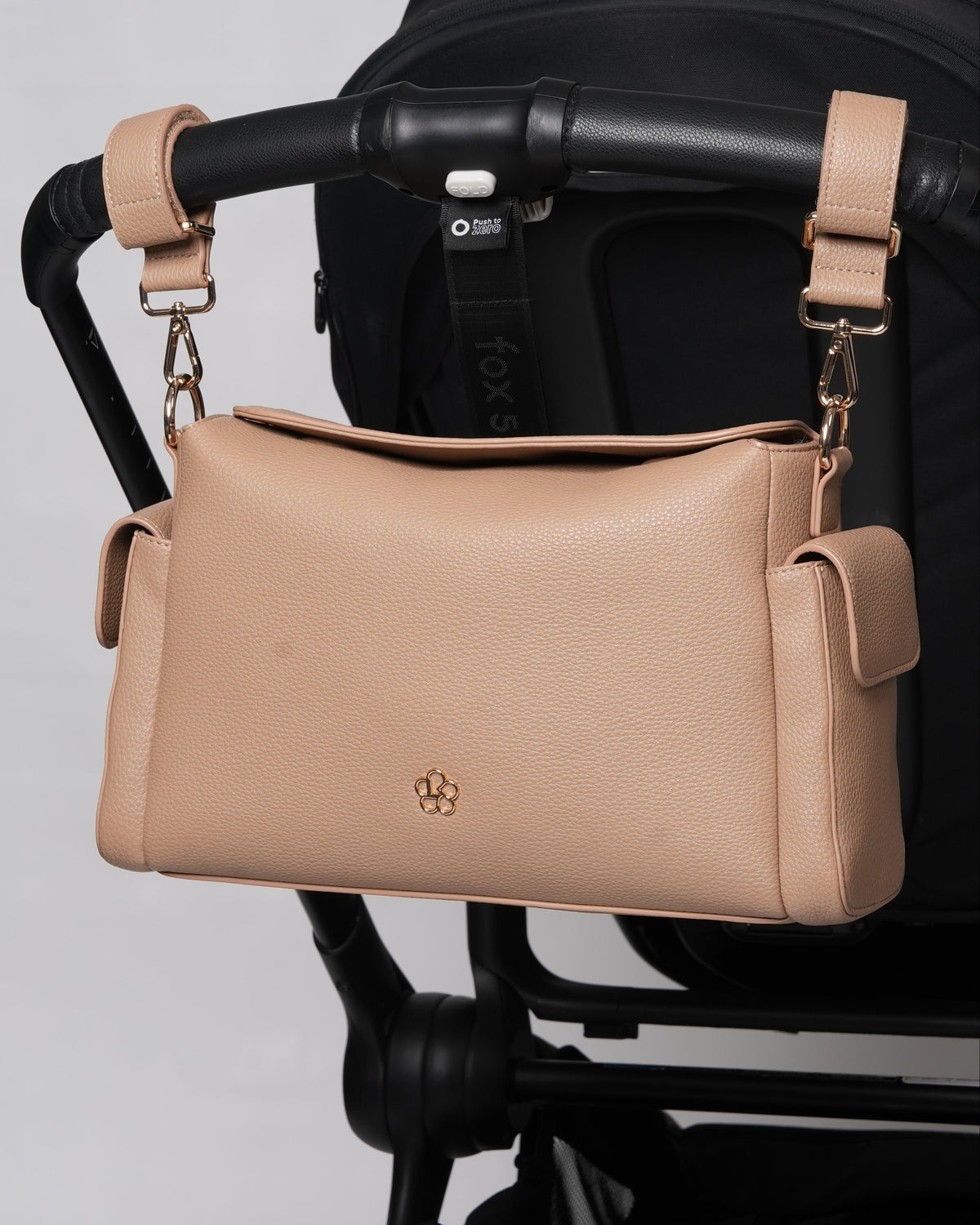 A beige pebbled leather crossbody bag with gold hardware and a small gold logo, hanging from a black stroller handlebar against a light gray background.