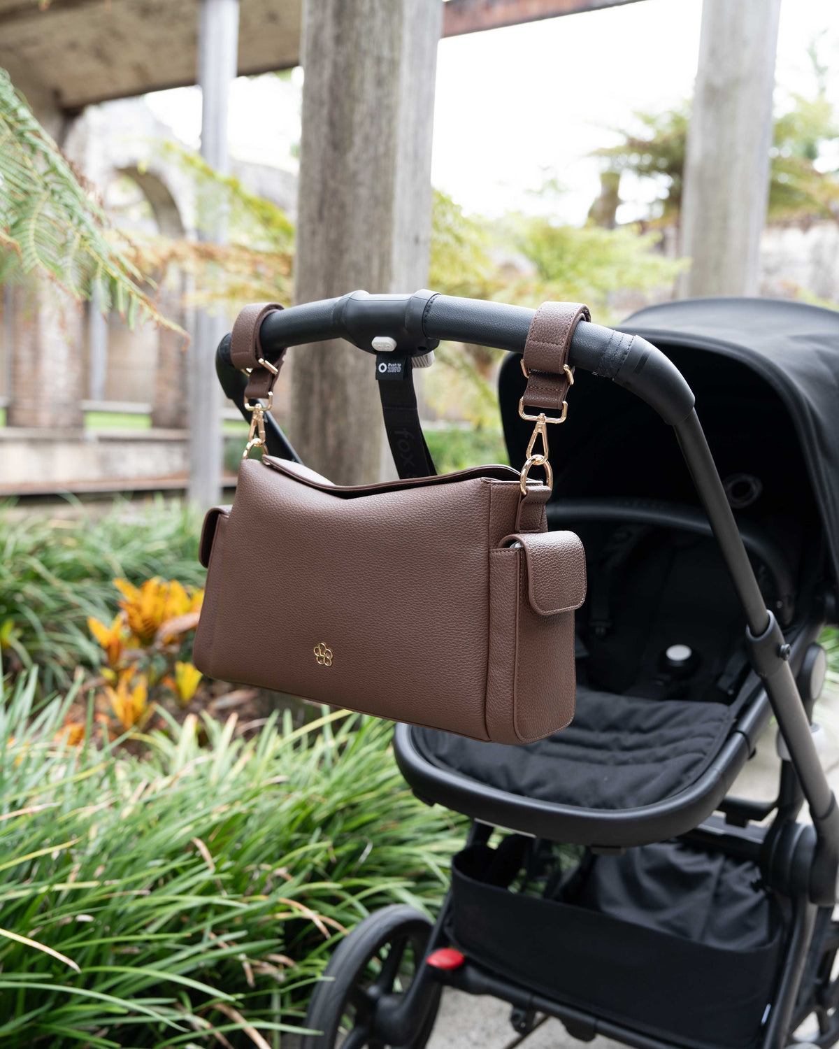 Lenelly Hera pram caddy bag in brown vegan leather with adjustable strap, hanging on a black stroller outdoors