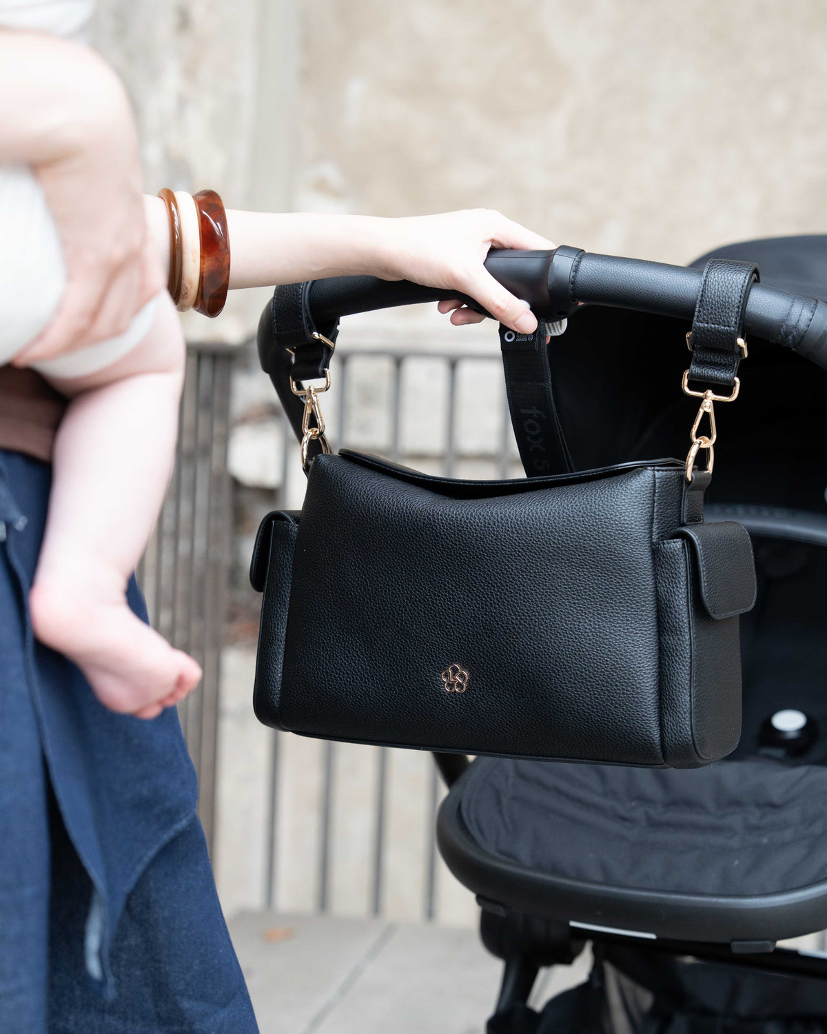 Lenelly Hera pram caddy bag in black vegan leather with adjustable strap, front view attached to a stroller with a woman holding a baby off frame and her hand on a stroller pram