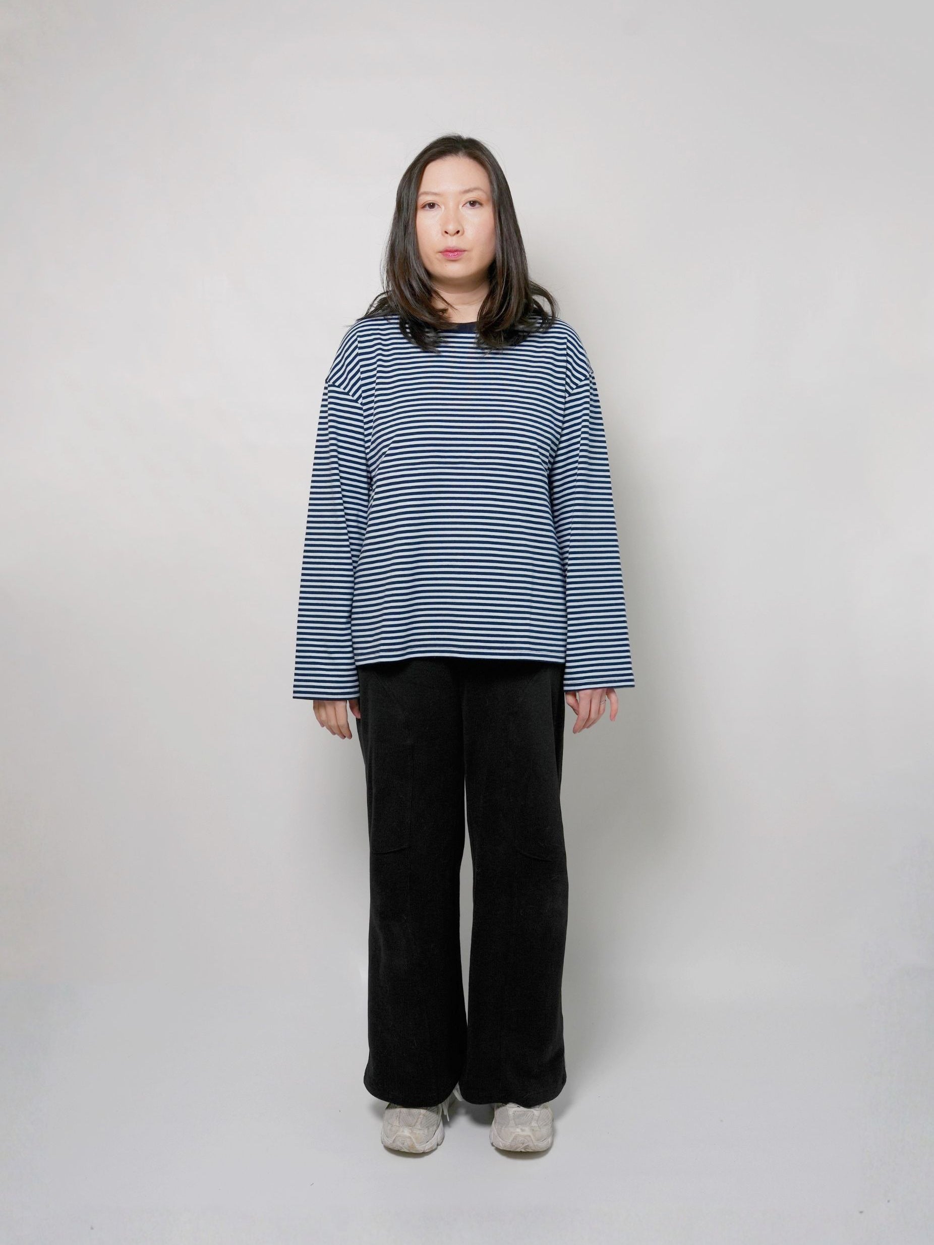 A woman stands against a plain light gray studio background wearing a loose long-sleeve top with thin sky blue and navy horizontal stripes, black wide-leg pants, and light gray shoes.