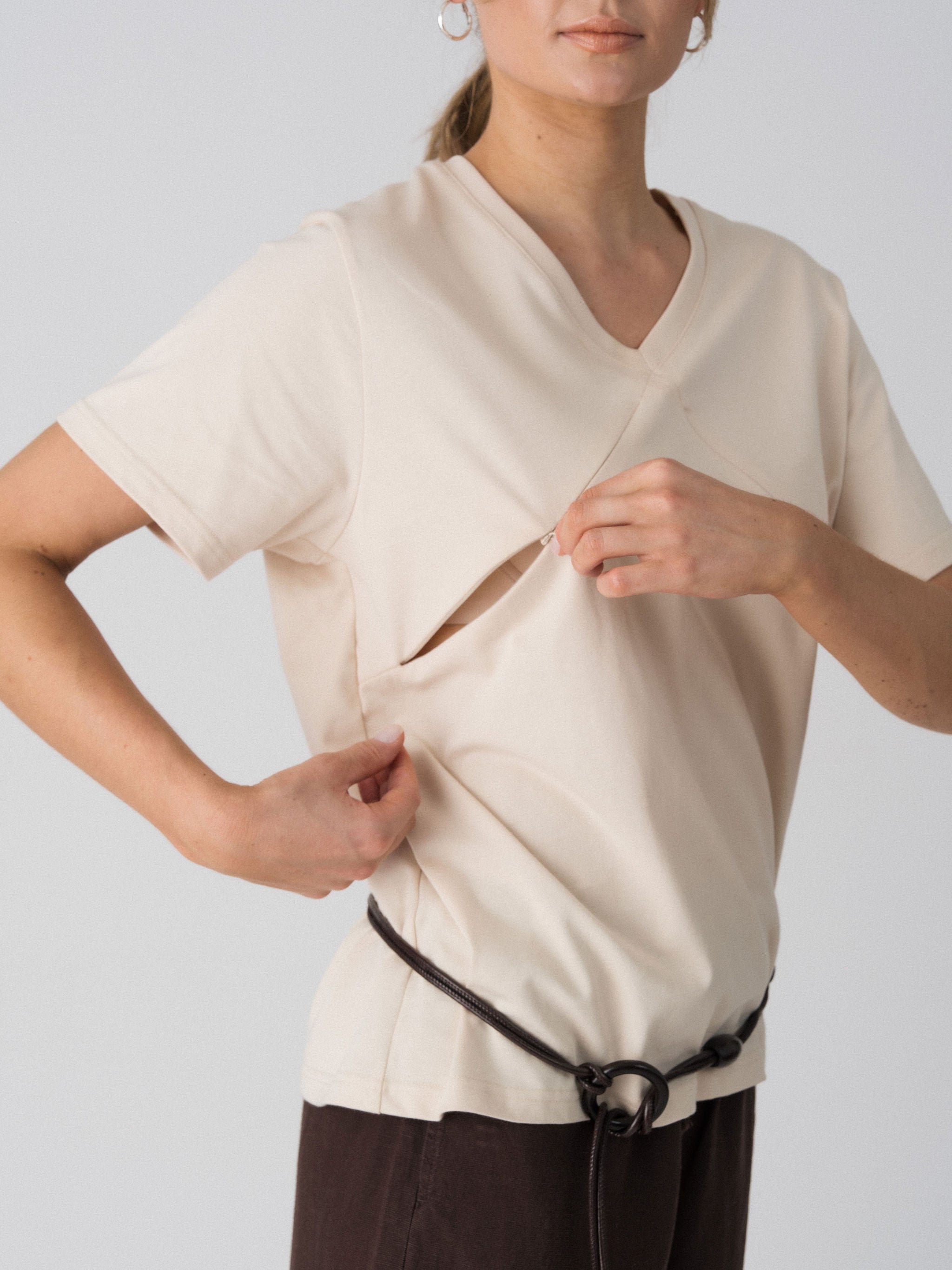 Model opening the hidden diagonal zip below the V-neckline of the Lenelly Avery tee in sand for discreet breastfeeding access