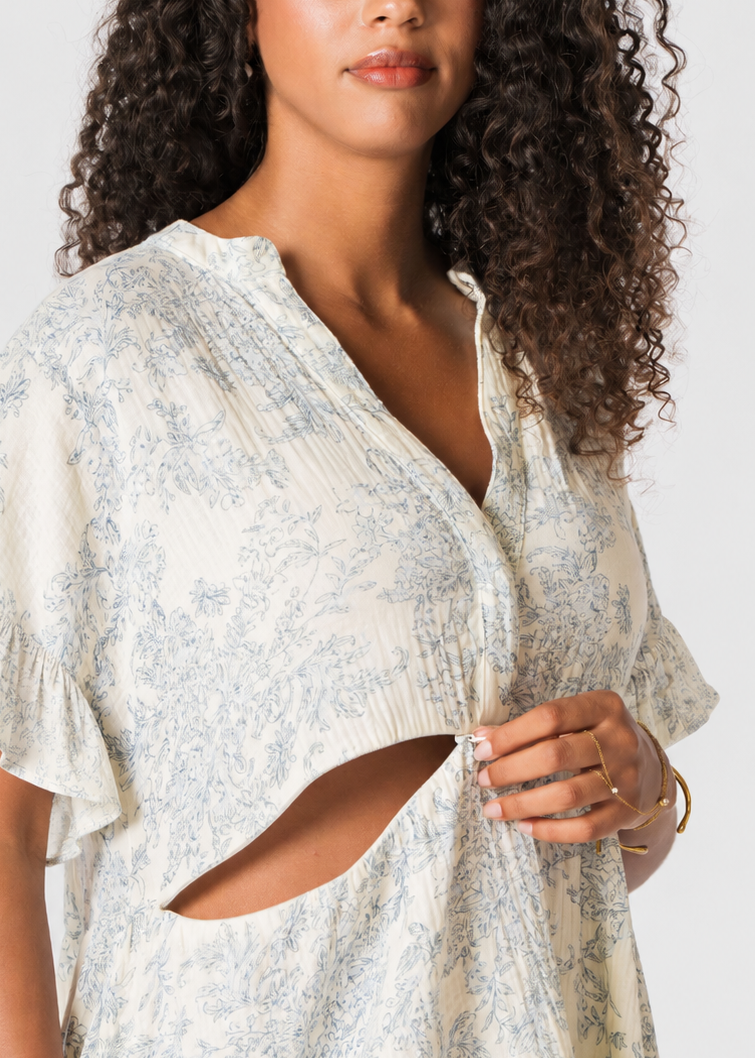 A woman wearing a light cream-colored short-sleeve tunic dress with a blue floral toile pattern, featuring a V-neckline and a distinctive side cutout, photographed against a neutral light gray background.