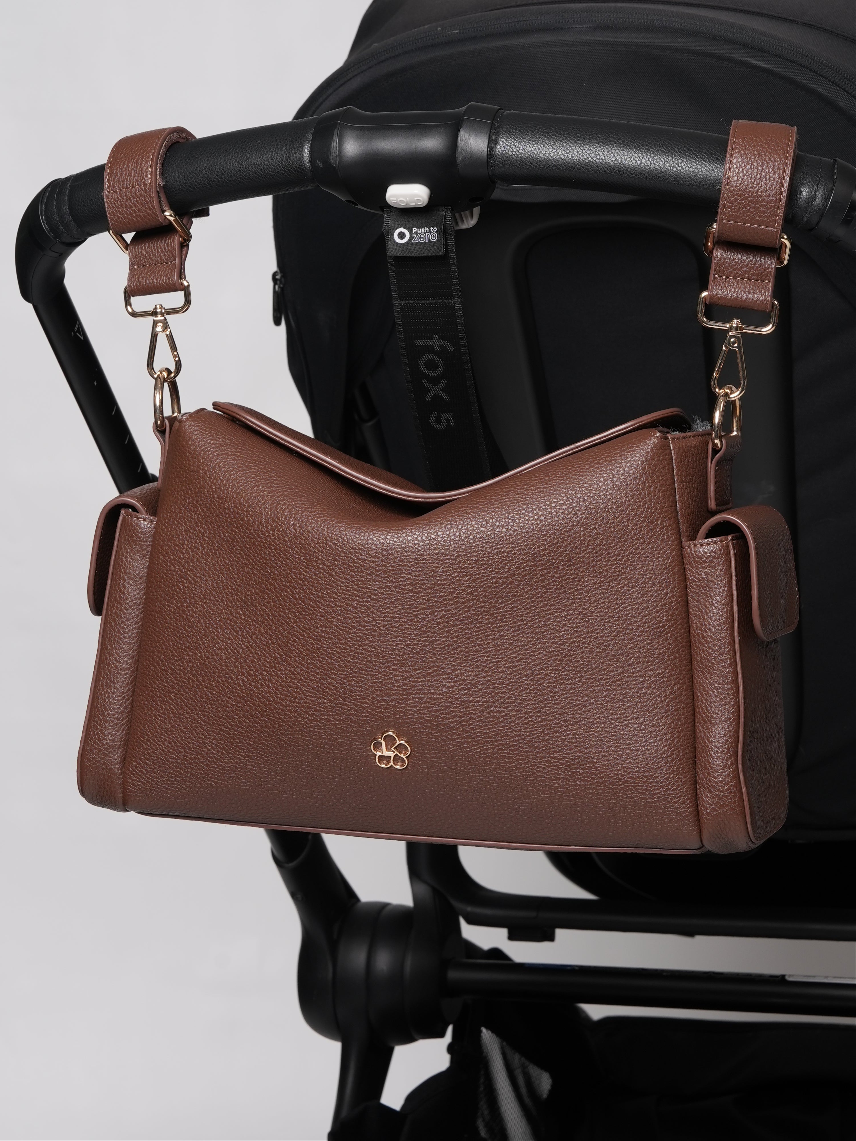 A brown pebbled leather shoulder bag with gold hardware and a small gold logo, hanging from a black stroller handlebar against a light gray background.