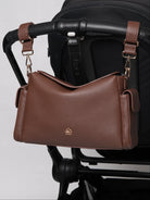 A brown pebbled leather shoulder bag with gold hardware and a small gold logo, hanging from a black stroller handlebar against a light gray background.