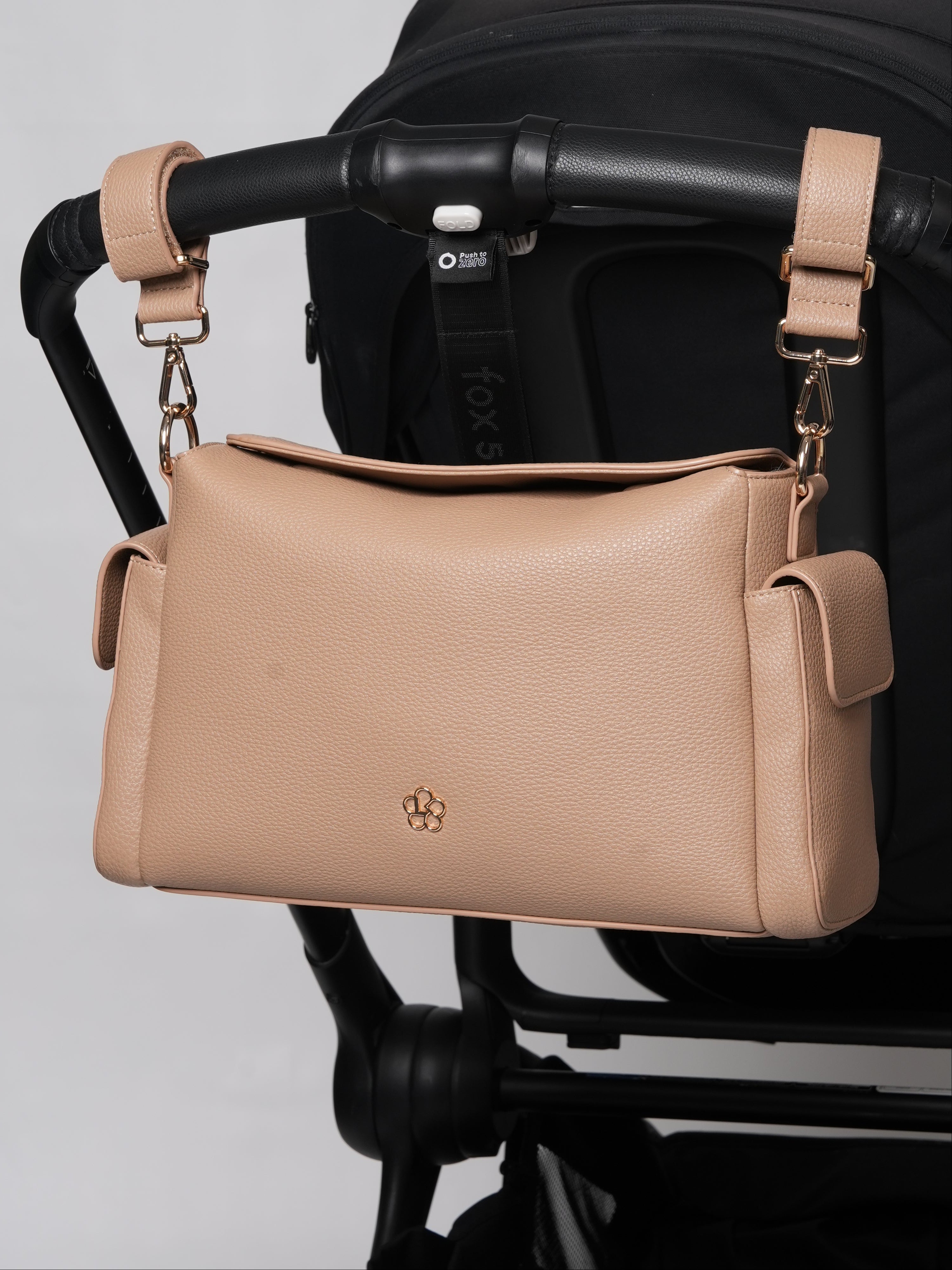 A beige pebbled leather crossbody bag with gold hardware and a small gold logo, hanging from a black stroller handlebar against a light gray background.
