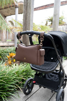 Lenelly Hera pram caddy bag in brown vegan leather with adjustable strap, hanging on a black stroller outdoors