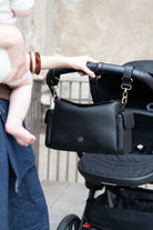 Lenelly Hera pram caddy bag in black vegan leather with adjustable strap, front view attached to a stroller with a woman holding a baby off frame and her hand on a stroller pram