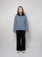 A woman stands against a plain light gray studio background wearing a loose long-sleeve top with thin sky blue and navy horizontal stripes, black wide-leg pants, and light gray shoes.